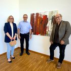 Drei Personen posieren vor einem Gemälde für ein Foto. Links steht eine Frau mit dunkelblauem Blazer, in der Mitte ein Mann mit einem hellblauen Hemd und rechts ein Mann mit einem grauen Sakko.