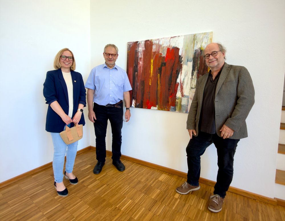 „Kunst und Kultur sind ein starkes Mittel für ein offenes Miteinander“ Drei Personen posieren vor einem Gemälde für ein Foto. Links steht eine Frau mit dunkelblauem Blazer, in der Mitte ein Mann mit einem hellblauen Hemd und rechts ein Mann mit einem grauen Sakko.