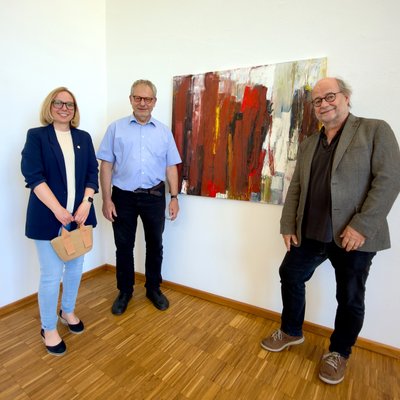 Drei Personen posieren vor einem Gemälde für ein Foto. Links steht eine Frau mit dunkelblauem Blazer, in der Mitte ein Mann mit einem hellblauen Hemd und rechts ein Mann mit einem grauen Sakko.