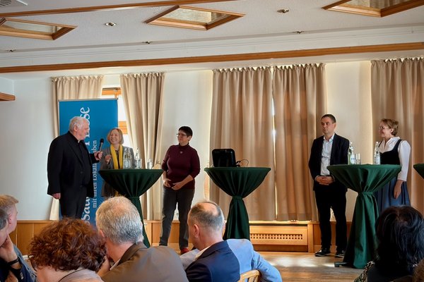 Fünf Personen stehen auf einer Bühne an drei Bistrotischen. Einer Person wird gerade das Mikrofon gereicht.