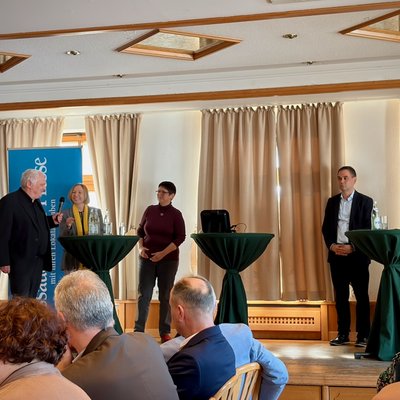 Fünf Personen stehen auf einer Bühne an drei Bistrotischen. Einer Person wird gerade das Mikrofon gereicht.