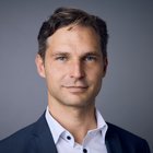 Ein Mann schaut in die Kamera. Er trägt ein dunkles Sakko und ein weißes Hemd. Seine Haare sind dunkelblond und kurz.