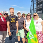 Sechs Personen sehen in der Sonne. Drei Personen tragen eine Mütze. Eine Person trägt eine grüne Fahne.