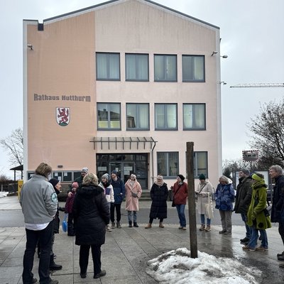 Etwa 15 Personen stehen vor dem Rathaus in Hutthurm. Sie hören einem Vortrag zu.
