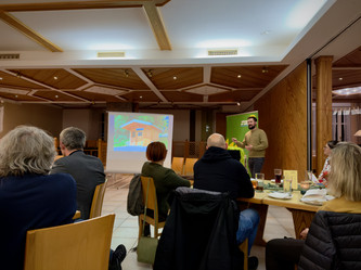 Ein Mann hält einen Vortrag vor einer Gruppe in einem Gastraum mit Holzvertäfelung. Auf der Leinwand hinter ihm ist eine Präsentation mit einem Haus zu sehen. Die Zuhörenden sitzen an Tischen mit Getränken und Unterlagen, einige machen sich Notizen.