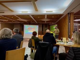 Ein Mann hält einen Vortrag vor einer Gruppe in einem Gastraum mit Holzvertäfelung. Auf der Leinwand hinter ihm ist eine Präsentation mit einem Haus zu sehen. Die Zuhörenden sitzen an Tischen mit Getränken und Unterlagen, einige machen sich Notizen.