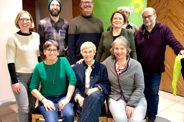 Neun Personen, drei sitzend und sechs stehend, lächeln in die Kamera bei einer Veranstaltung mit grünem Banner im Hintergrund.