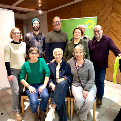 Neun Personen, drei sitzend und sechs stehend, lächeln in die Kamera bei einer Veranstaltung mit grünem Banner im Hintergrund.