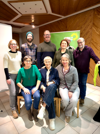 Neun Personen, drei sitzend und sechs stehend, lächeln in die Kamera bei einer Veranstaltung mit grünem Banner im Hintergrund.