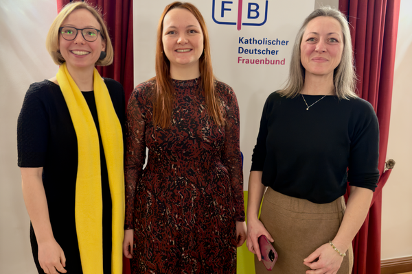 Drei Frauen posieren für ein Foto. Sie lächeln in die Kamera. Die Frauen tragen festliche Kleidung. Hinter den Frauen steht ein Roll-up mit dem Text "KDFB".