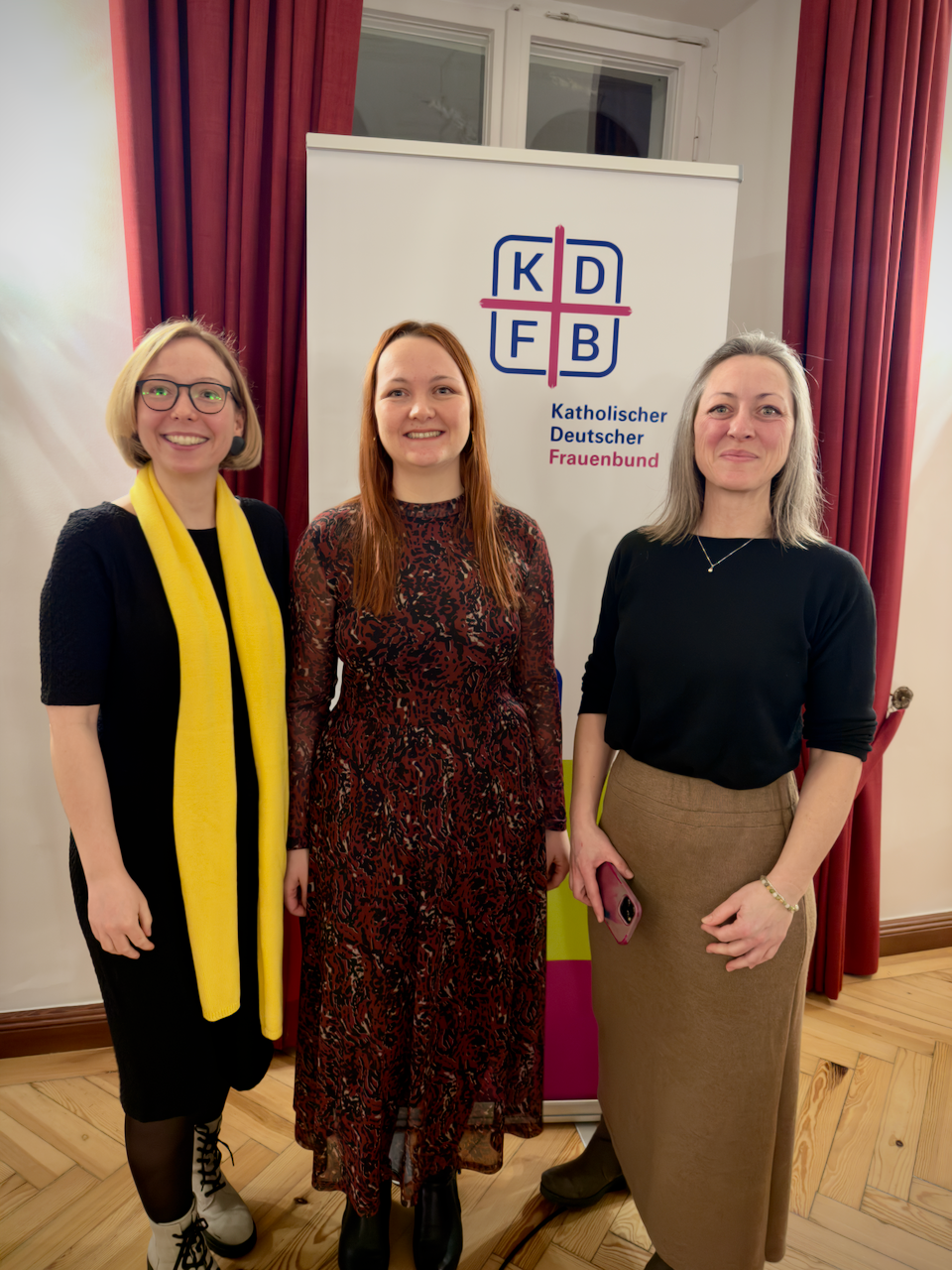 Drei Frauen posieren für ein Foto. Sie lächeln in die Kamera. Die Frauen tragen festliche Kleidung. Hinter den Frauen steht ein Roll-up mit dem Text "KDFB".