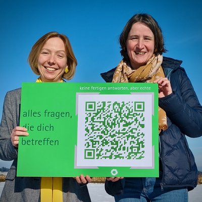 Zwei Frauen halten eine Postkarte im XXL-Format in den Händen. Auf der Postkarte steht "alles fragen, die dich betreffen"