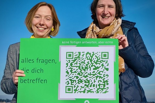 Zwei Frauen halten eine Postkarte im XXL-Format in den Händen. Auf der Postkarte steht "alles fragen, die dich betreffen"