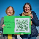 Zwei Frauen halten eine Postkarte im XXL-Format in den Händen. Auf der Postkarte steht "alles fragen, die dich betreffen"