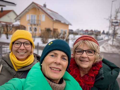 Drei Frauen lächeln in die Kamera. Es ist Winter. Sie tragen dicke Jacken und eine gelbe, grüne sowie rote Mütze.