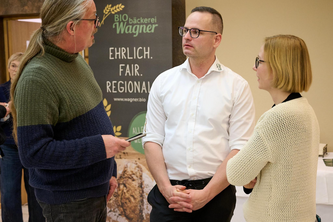 Drei Personen stehen vor einem Banner der Bio-Bäckerei Wagner und diskutieren, ein Mann hält einen Stift in der Hand. Im Hintergrund hängt ein Banner mit dem Logo der Bio-Bäckerei Wagner und den Worten EHRLICH. FAIR. REGIONAL! sowie der Webadresse www.wagner.bio.
