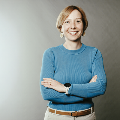 Eine Frau mit blauem Pullover lächelt in die Kamera. Sie hat kurze blonde Haare und verschränkt die Arme.
