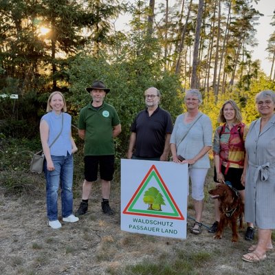 Sechs Personen stehen in einer Lichtung im Wald. Vor ihnen steht eine Tafel mit der Aufschrift "Waldschutz Passauer Land".