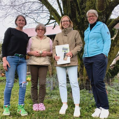 Vier Frauen lächeln in die Kamera. Sie stehen in einem Garten. Sie tragen Pullover und Jacken, es ist Frühling.