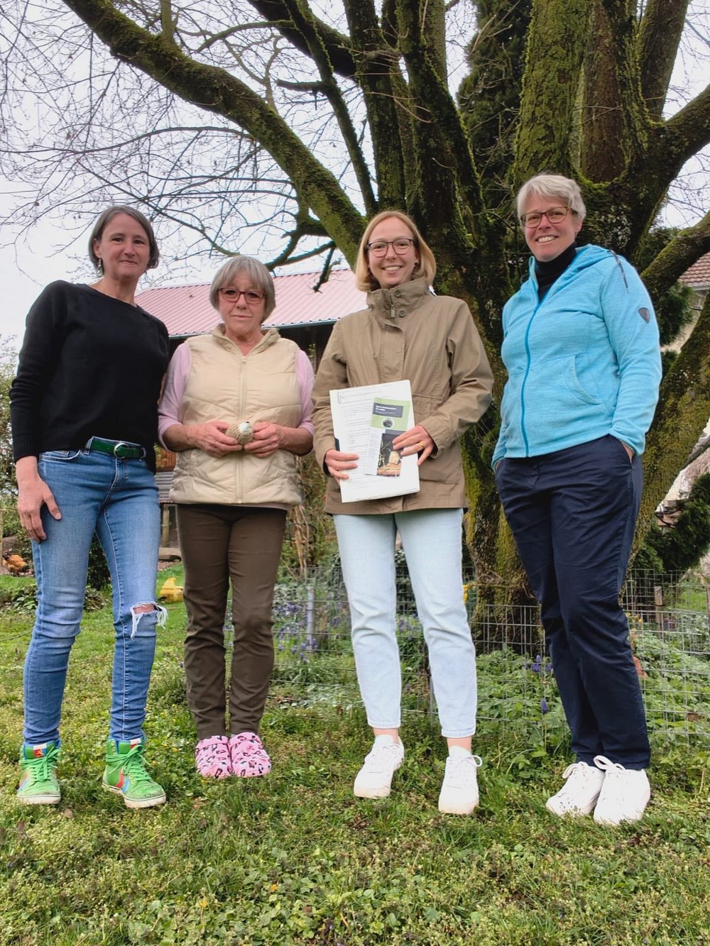 Vier Frauen lächeln in die Kamera. Sie stehen in einem Garten. Sie tragen Pullover und Jacken, es ist Frühling.