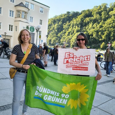 Zwei Frauen halten eine Fahne und ein Plakat. Auf dem Plakat steht "Es reicht. Das Leben bezahlbar machen. Niederbayern". Die Fahne ist grün und darauf steht "Bündnis 90 / Die Grünen".