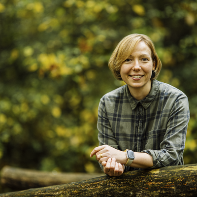 Eine Frau mit kurzen blonden Haaren stützt ihre Arme auf ein Brückengeländer. Sie trägt ein grünes Hemd und lächelt. Die Frau ist im Wald auf einer Holzbrücke.