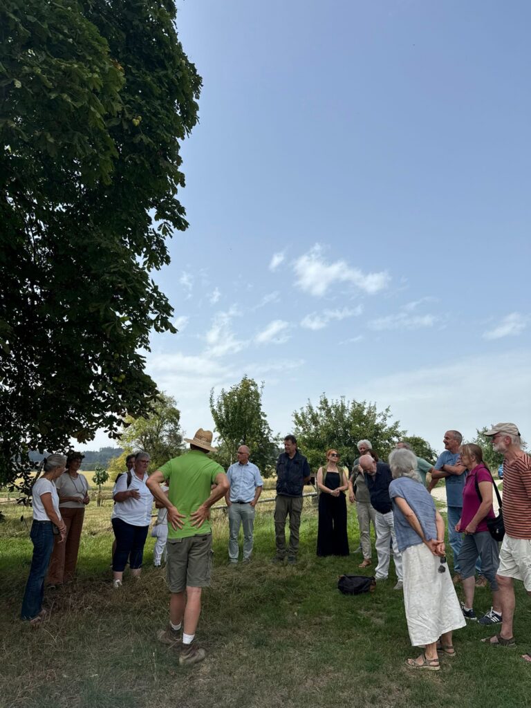 Zirka zwanzig Personen lauschen einem Vortrag auf einer Wiese. Die Sonne scheint.