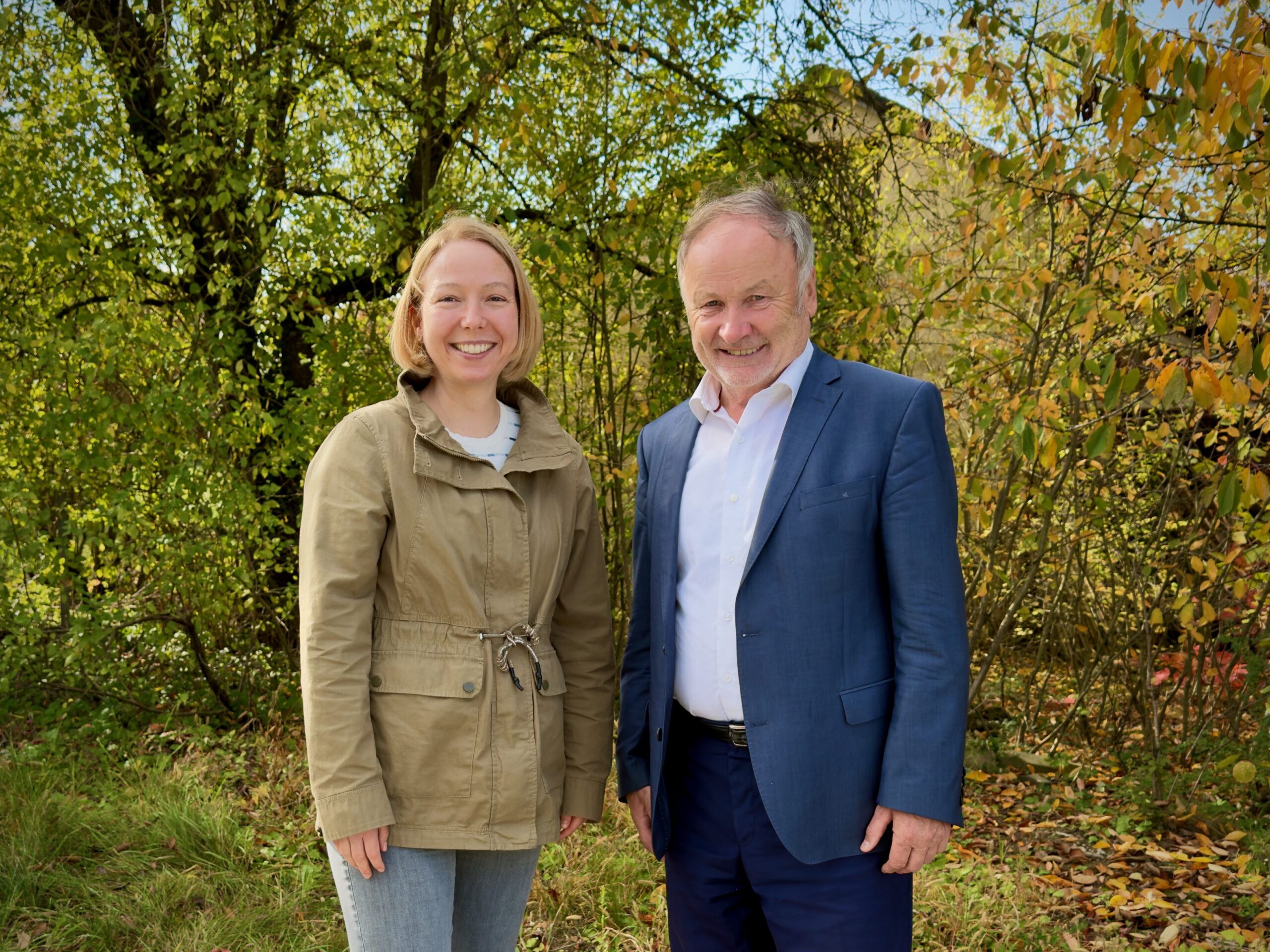 Zwei Personen stehen vor Sträuchern. Es ist Herbst. Rechts steht ein Mann mit weißem Hemd und blauem Sakko. Links steht eine Frau mit weißem Pullover und grüner Jacke.